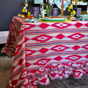 Peut inclure: Une nappe blanche avec un motif ikat rose et jaune et un bord festonn&eacute;. La nappe est dress&eacute;e avec des assiettes, des verres et des chandeliers.