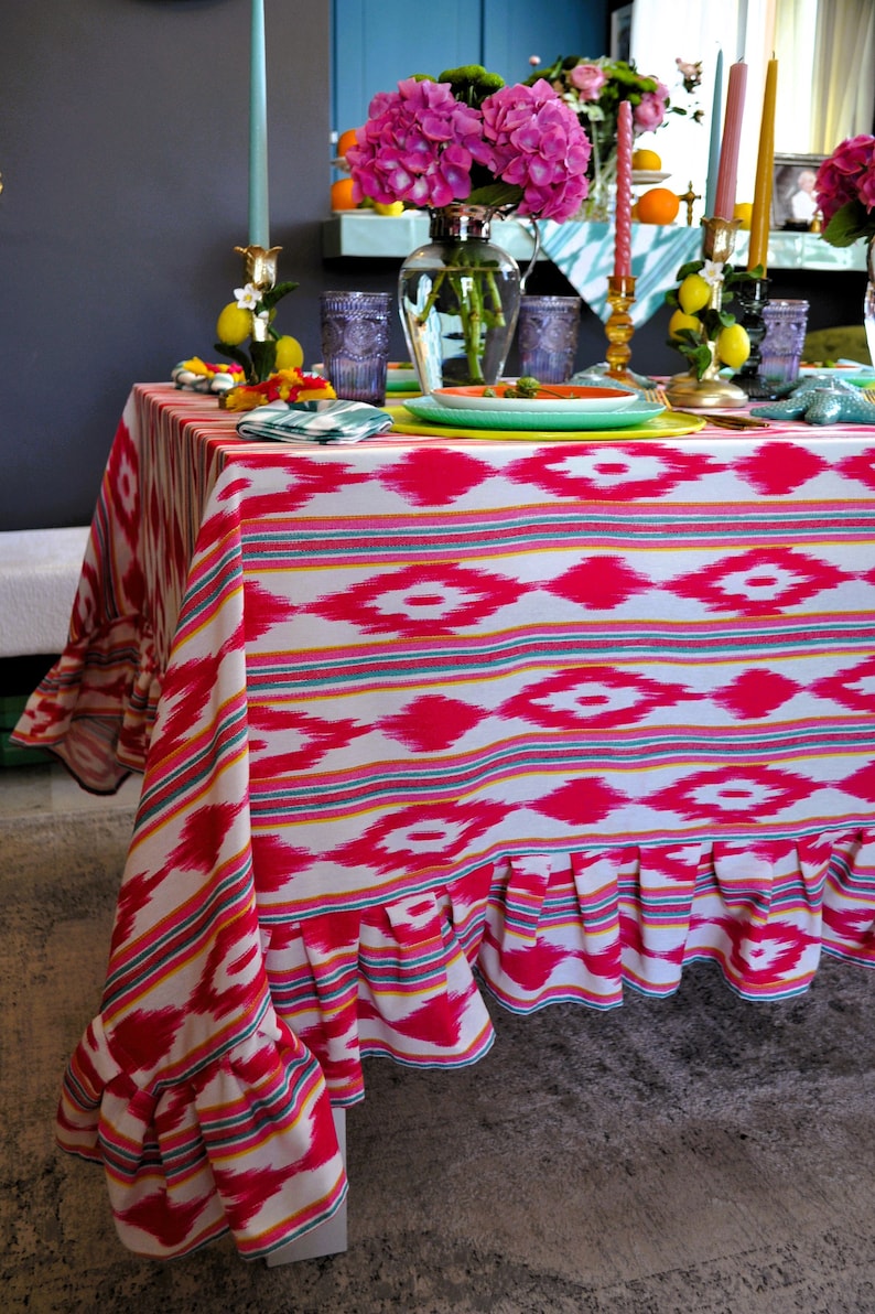 Peut inclure: Une nappe blanche avec un motif ikat rose et jaune et un bord festonn&eacute;. La nappe est dress&eacute;e avec des assiettes, des verres et des chandeliers.
