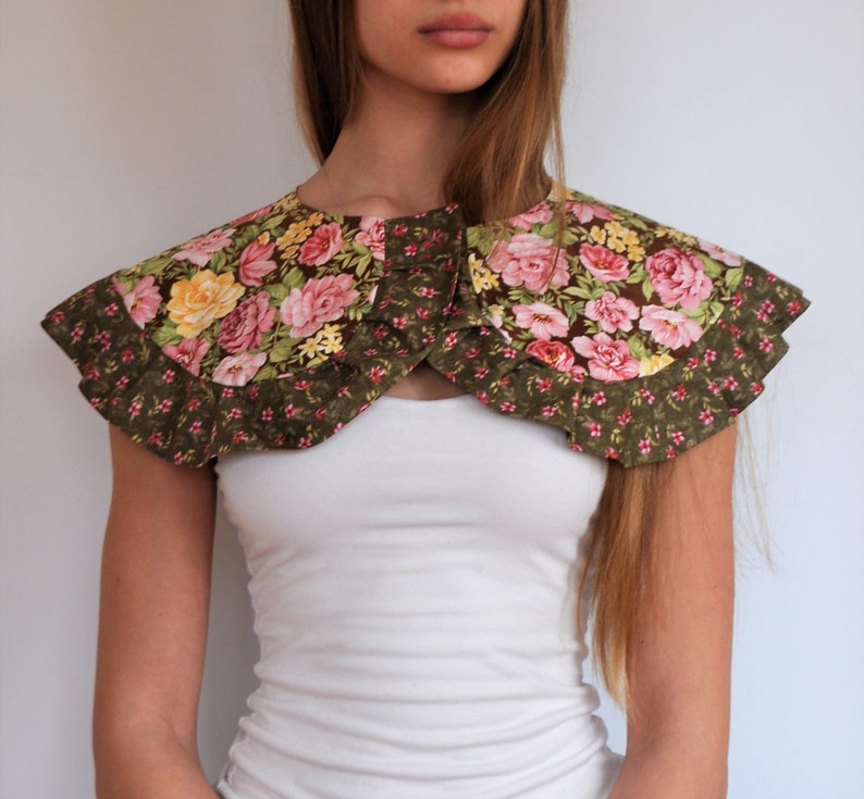 May include: A green and pink floral fabric collar with ruffled edges. The collar is worn over a white shirt.