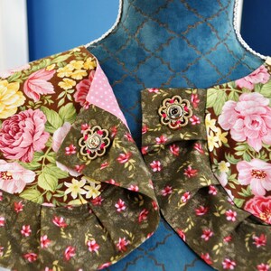 May include: A brown and green floral fabric collar with pink and yellow flowers. The collar has two gold-toned buttons with a decorative design.