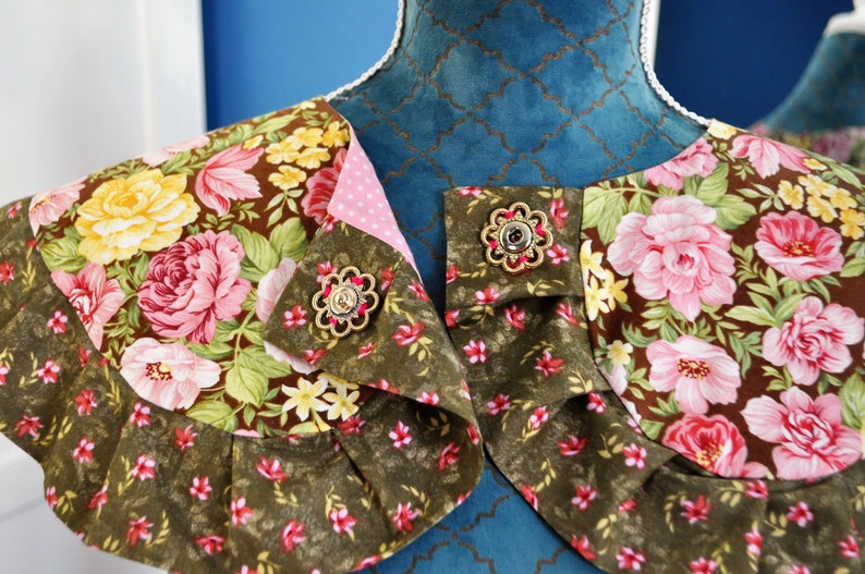 May include: A brown and green floral fabric collar with pink and yellow flowers. The collar has two gold-toned buttons with a decorative design.