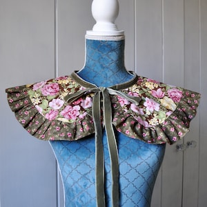 May include: A green and pink floral fabric collar with a green velvet bow tie. The collar is displayed on a blue velvet mannequin.