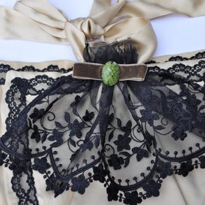 May include: A beige satin garment with black floral lace trim and a decorative bow. A brown velvet ribbon and a green oval jewel accent the design. The garment has a satin ribbon tie.