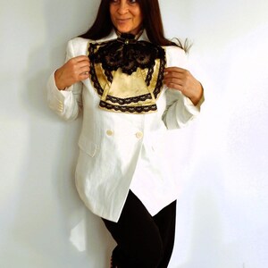 May include: A white blazer with a double-breasted design, paired with black pants. The blazer features a decorative gold and black lace collar. The outfit is completed with leopard print shoes, creating a contrast in textures and patterns.