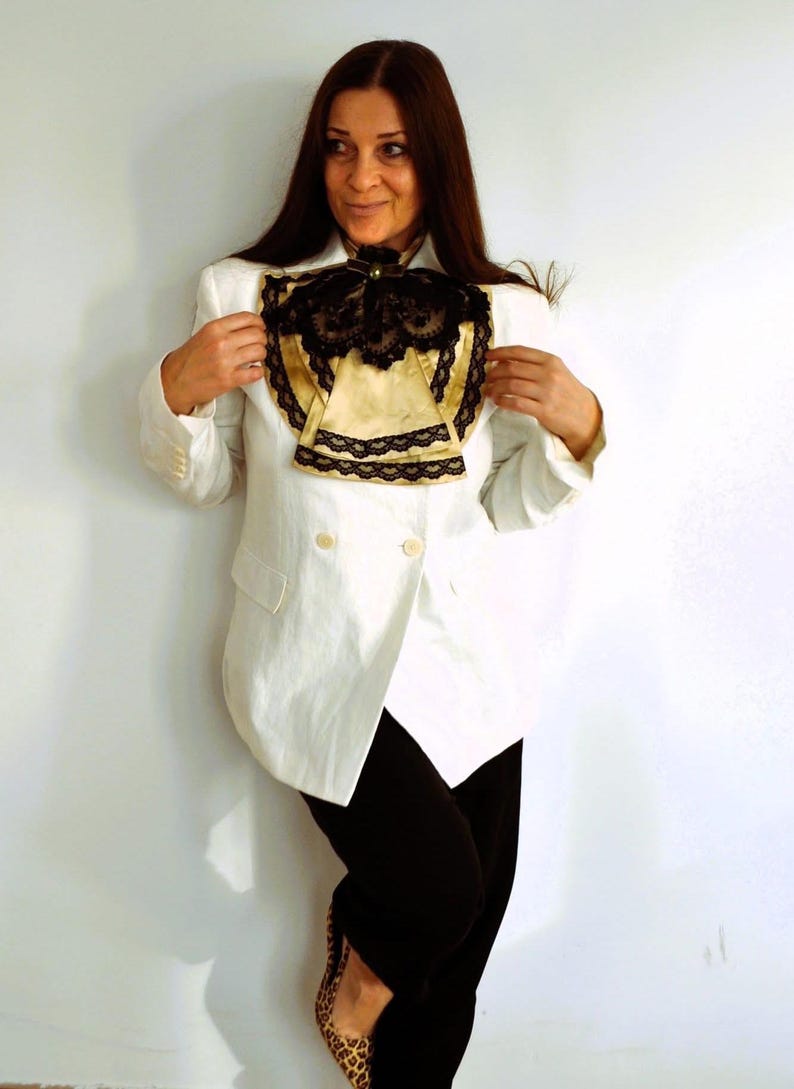 May include: A white blazer with a double-breasted design, paired with black pants. The blazer features a decorative gold and black lace collar. The outfit is completed with leopard print shoes, creating a contrast in textures and patterns.