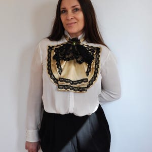 May include: A white blouse with a gold satin ascot and black lace trim. The ascot is adorned with a black bow and a green jewel. The blouse is paired with black pants.