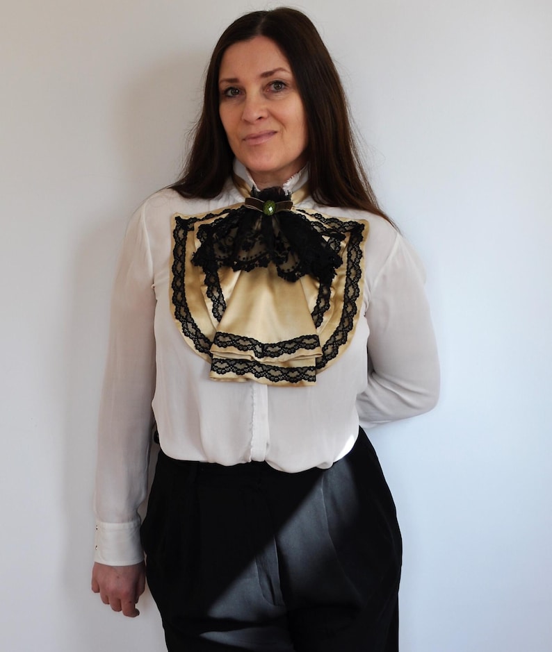 May include: A white blouse with a gold satin ascot and black lace trim. The ascot is adorned with a black bow and a green jewel. The blouse is paired with black pants.