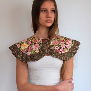 May include: A green floral fabric collar with pink and yellow flowers. The collar has a ruffled edge and is worn over a white tank top.