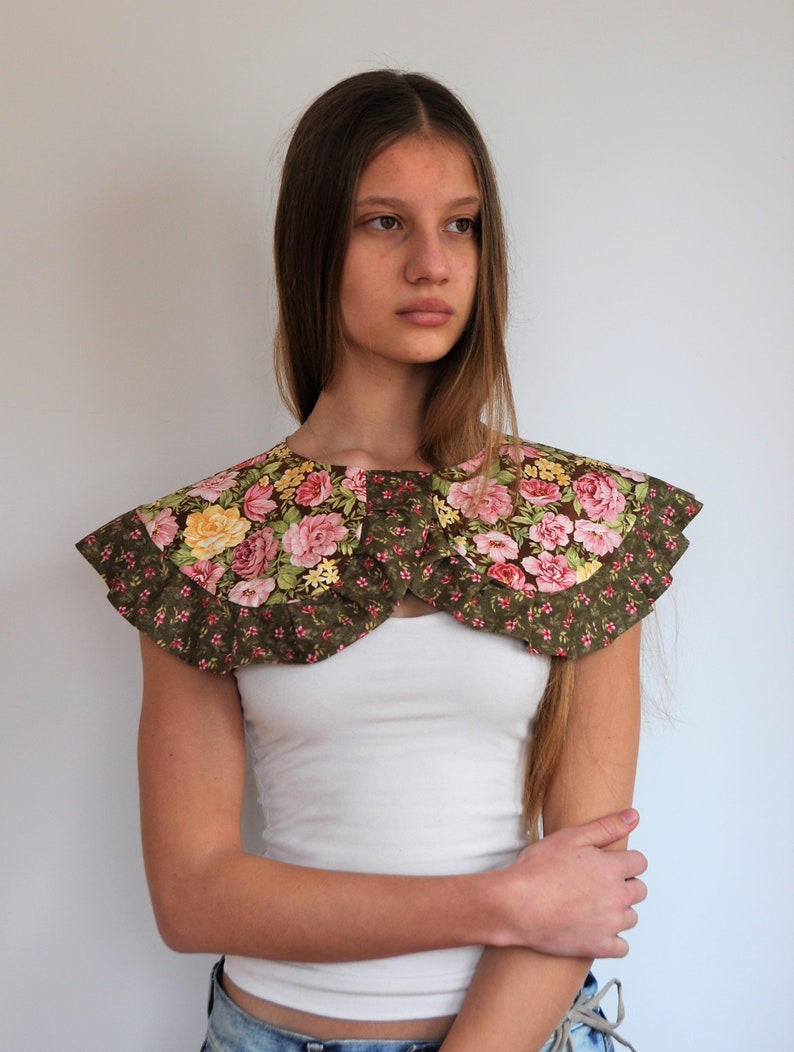 May include: A green floral fabric collar with pink and yellow flowers. The collar has a ruffled edge and is worn over a white tank top.