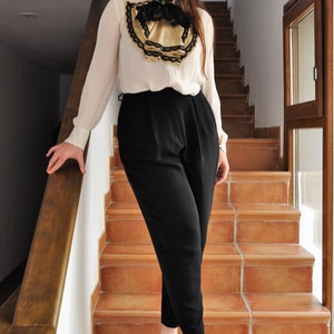 May include: A woman's outfit featuring a white blouse with a black and gold decorative collar, paired with black trousers and beige heels with black caps. The outfit is worn on a staircase with brown tile steps.