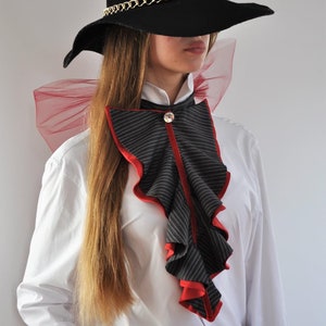 May include: A white button-down shirt with a black and red ruffled collar. The collar has a red velvet trim and a rhinestone button. The person is wearing a black wide-brimmed hat with a gold chain.