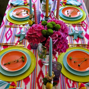 Peut inclure: Une table dress&eacute;e de mani&egrave;re color&eacute;e avec une composition florale rose et verte au centre, des assiettes orange, des assiettes de pr&eacute;sentation bleues et des couverts dor&eacute;s. La table est recouverte d'une nappe ikat blanche et rose.