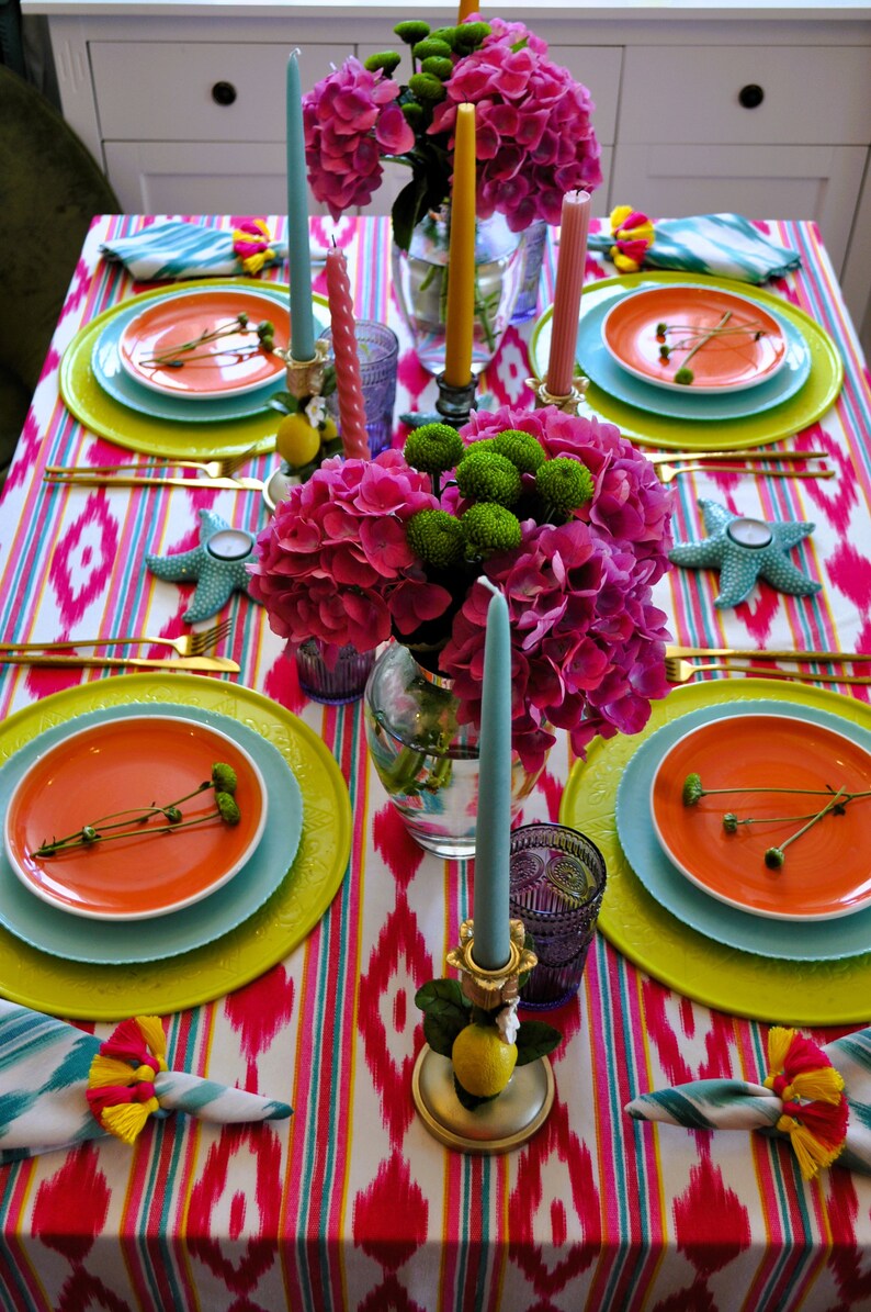Peut inclure: Une table dress&eacute;e de mani&egrave;re color&eacute;e avec une composition florale rose et verte au centre, des assiettes orange, des assiettes de pr&eacute;sentation bleues et des couverts dor&eacute;s. La table est recouverte d'une nappe ikat blanche et rose.