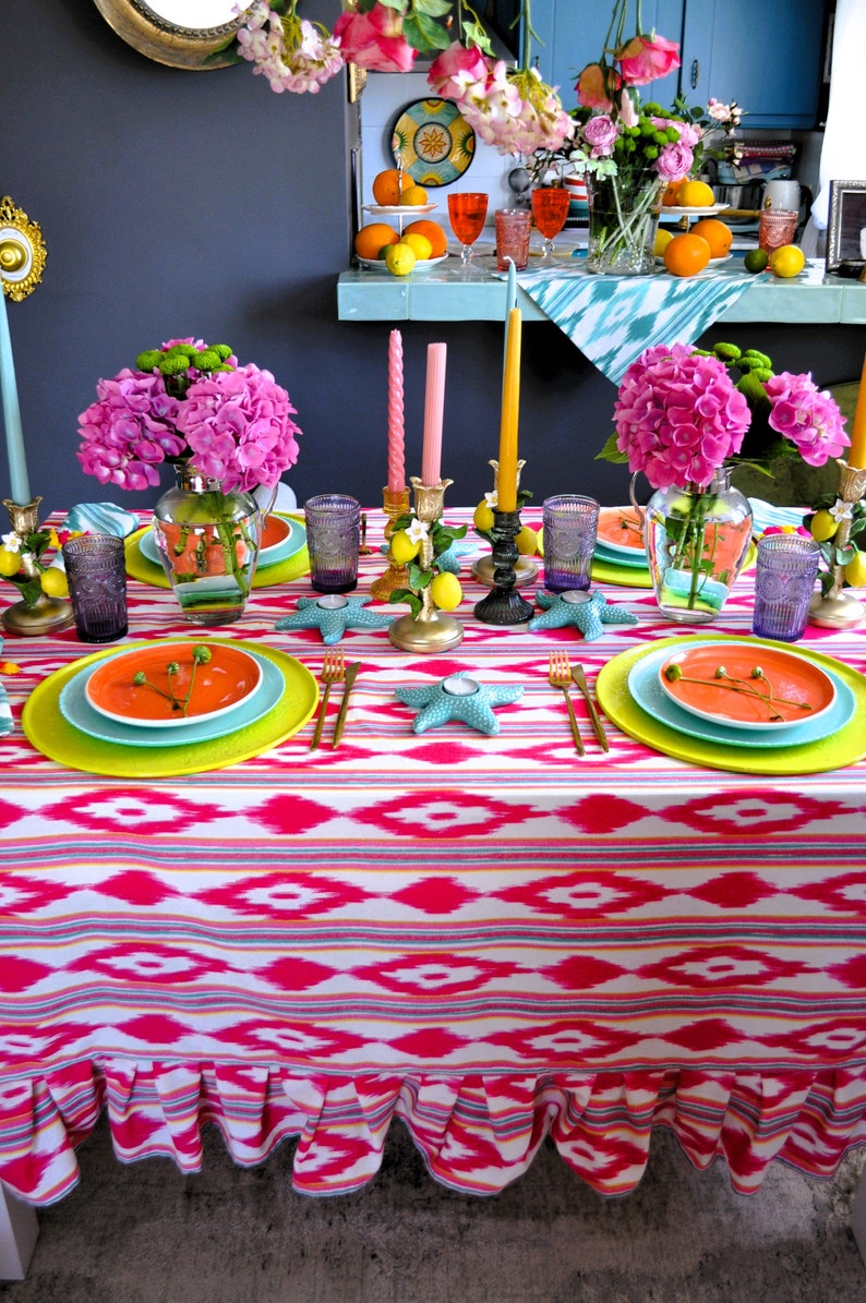 Peut inclure: Une nappe rose et blanche avec un motif g&eacute;om&eacute;trique et un bord festonn&eacute;. La nappe est dress&eacute;e pour un d&icirc;ner avec des assiettes, des couverts, des bougies et des fleurs.