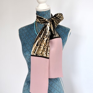 Peut inclure: Un foulard à motif rose et marron noué autour du cou d'un mannequin bleu. Le foulard a un motif peau de serpent et une section rose unie.