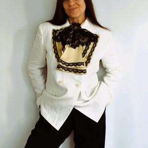 May include: A white double-breasted blazer with black pants. The blazer features a decorative cream-colored ascot with black lace trim. The outfit is completed with black pants and a dark green brooch.
