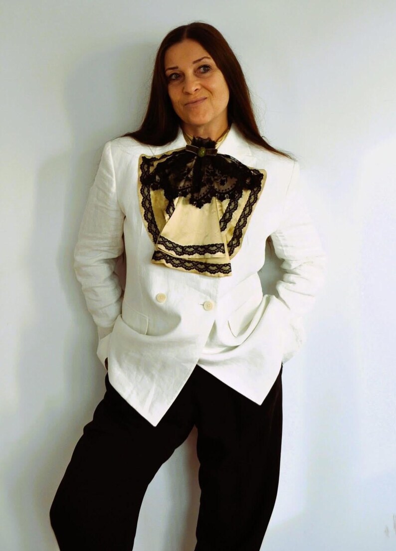 May include: A white double-breasted blazer with black pants. The blazer features a decorative cream-colored ascot with black lace trim. The outfit is completed with black pants and a dark green brooch.