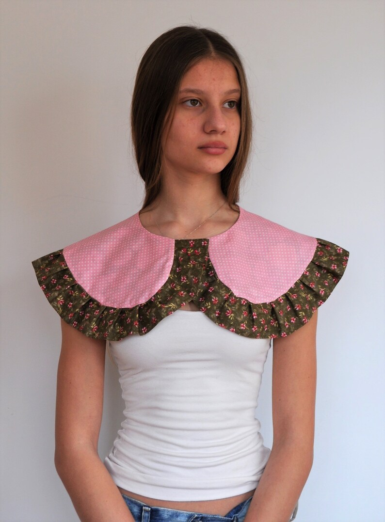 May include: A pink and green fabric collar with a ruffled edge. The collar is worn over a white shirt.