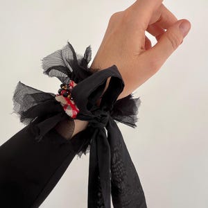 Puede incluir: Puño de muñeca negro con un diseño en capas y con volantes. El puño presenta un lazo y una pequeña sección con tela de cuadros rojos y blancos y cuentas. El puño está hecho de tela transparente y fluida.
