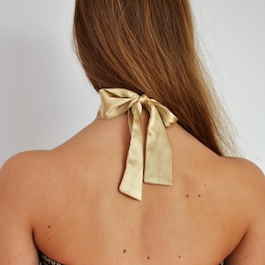 May include: Back view of a person wearing a gold satin halter top with a bow detail at the neck. The top is black and gold patterned. Long, light brown hair cascades down the back.