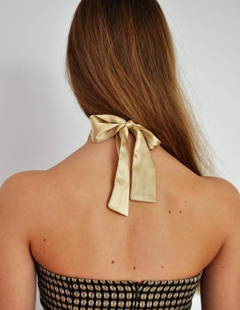 May include: Back view of a person wearing a gold satin halter top with a bow detail at the neck. The top is black and gold patterned. Long, light brown hair cascades down the back.