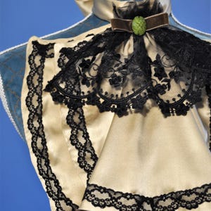 May include: A cream-colored satin collar with black lace trim and a black lace overlay. A brown velvet ribbon and a green jewel accent the collar. The collar is displayed on a blue mannequin.