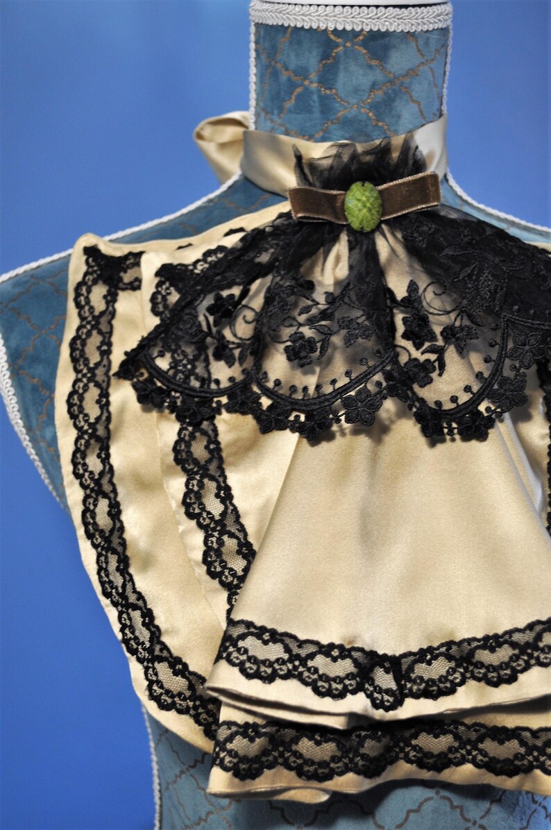 May include: A cream-colored satin collar with black lace trim and a black lace overlay. A brown velvet ribbon and a green jewel accent the collar. The collar is displayed on a blue mannequin.