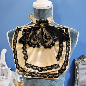 May include: A gold-colored satin neckpiece with black lace trim and a black lace overlay. The neckpiece is adorned with a brown velvet ribbon and a green jewel. The item is displayed on a blue mannequin.