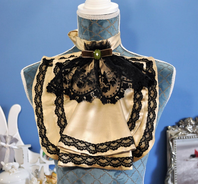 May include: A gold-colored satin neckpiece with black lace trim and a black lace overlay. The neckpiece is adorned with a brown velvet ribbon and a green jewel. The item is displayed on a blue mannequin.