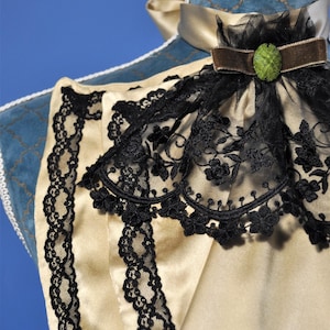 May include: Close-up of a cream-colored satin garment with black lace trim. The garment features a high collar with a black lace overlay and a brown velvet ribbon with a green jewel. The background is blue.