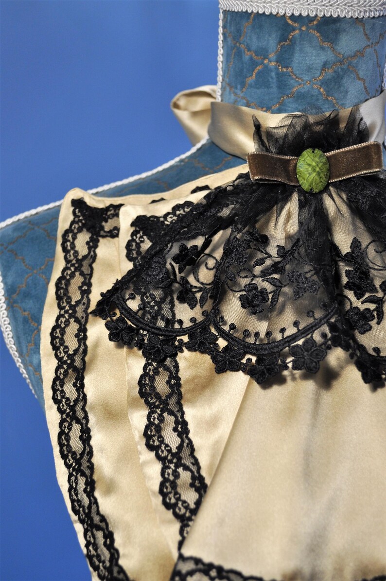 May include: Close-up of a cream-colored satin garment with black lace trim. The garment features a high collar with a black lace overlay and a brown velvet ribbon with a green jewel. The background is blue.