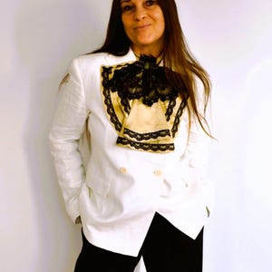 May include: A white blazer with a double-breasted design, paired with black pants. The blazer features a decorative cream-colored insert with black lace trim. The outfit is completed with a black bow.
