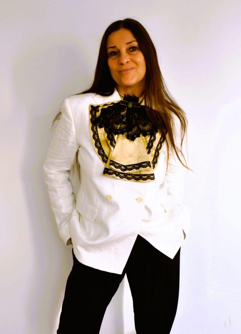 May include: A white blazer with a double-breasted design, paired with black pants. The blazer features a decorative cream-colored insert with black lace trim. The outfit is completed with a black bow.