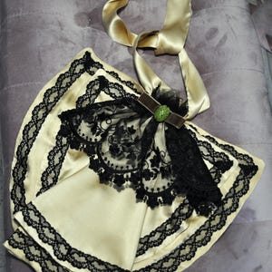 May include: A cream-colored, satin-like accessory with black lace trim and a black lace overlay. A gold satin ribbon is attached, along with a brown velvet accent and a green jewel. The item is likely a vintage-inspired fashion accessory.