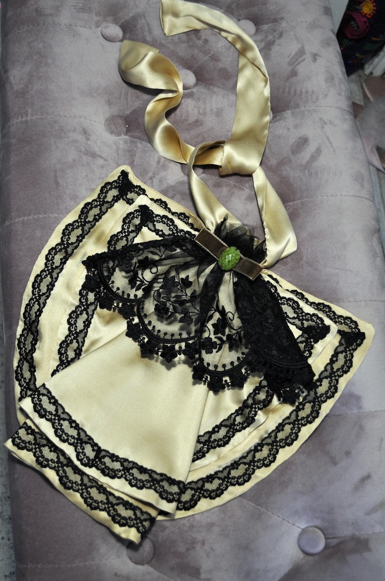 May include: A cream-colored, satin-like accessory with black lace trim and a black lace overlay. A gold satin ribbon is attached, along with a brown velvet accent and a green jewel. The item is likely a vintage-inspired fashion accessory.