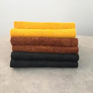 Peut inclure: Une pile de six pièces rectangulaires de matériau texturé disposées par paires de couleurs. Les deux du haut sont jaune vif, les deux du milieu sont brun-rouge foncé et les deux du bas sont noir uni. Chaque pièce a une surface fibreuse rugueuse.