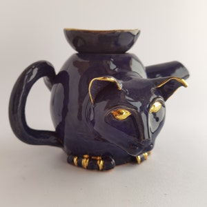 Op de afbeelding: Een donkerblauwe theepot van keramiek in de vorm van een kat met gouden accenten. De theepot heeft een deksel en een tuit. De ogen van de kat zijn goud.