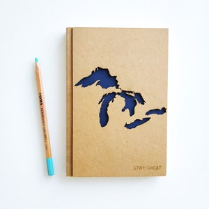 May include: A brown notebook with a blue cutout of the Great Lakes. The notebook has the text "STAY GREAT" printed on the front.