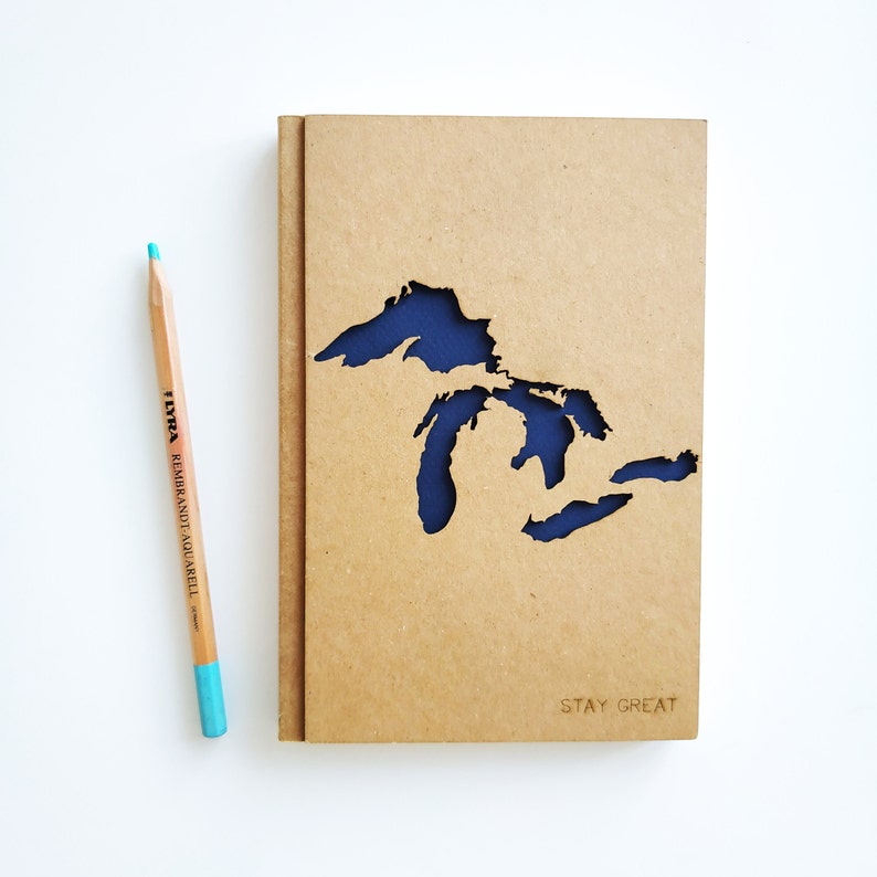 May include: A brown notebook with a blue cutout of the Great Lakes. The notebook has the text "STAY GREAT" printed on the front.