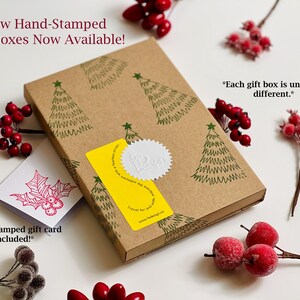 May include: A brown gift box with a hand-stamped design of Christmas trees. The box is closed and has a yellow sticker with a white seal. The text "New Hand-Stamped Gift Boxes Now Available!" is at the top of the image. The text "*Each gift box is uniquely different.*" is on the right side of the image. The text "*A hand-stamped gift card is included!*" is on the left side of the image.
