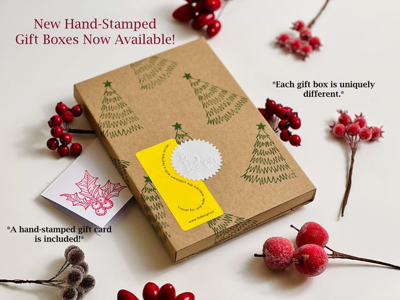 May include: A brown gift box with a hand-stamped design of Christmas trees. The box is closed and has a yellow sticker with a white seal. The text "New Hand-Stamped Gift Boxes Now Available!" is at the top of the image. The text "*Each gift box is uniquely different.*" is on the right side of the image. The text "*A hand-stamped gift card is included!*" is on the left side of the image.