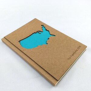 Puede incluir: Un cuaderno de cart&oacute;n marr&oacute;n con un recorte de los Estados Unidos en forma de mapa. El recorte est&aacute; relleno con un papel azul brillante. El cuaderno tiene el texto "The Paperbots 2016" impreso en el lomo.