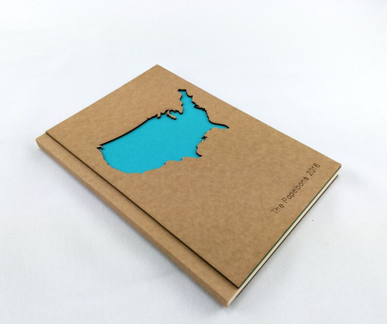 Puede incluir: Un cuaderno de cart&oacute;n marr&oacute;n con un recorte de los Estados Unidos en forma de mapa. El recorte est&aacute; relleno con un papel azul brillante. El cuaderno tiene el texto "The Paperbots 2016" impreso en el lomo.