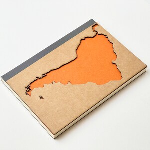 May include: A brown notebook with a grey spine. The cover has a cut-out shape of an island with an orange background.