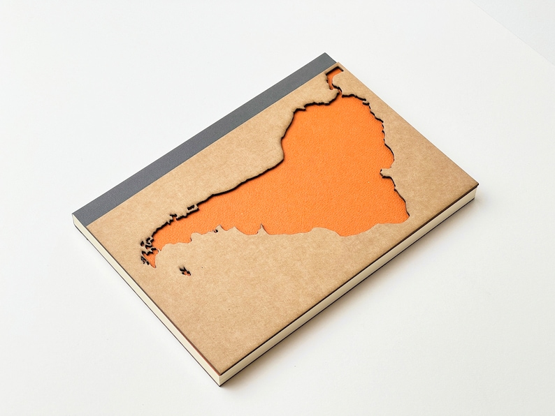 May include: A brown notebook with a grey spine. The cover has a cut-out shape of an island with an orange background.