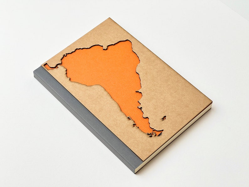 May include: A brown notebook with a cut-out shape of South America. The cut-out is filled with orange paper. The notebook has a grey spine.
