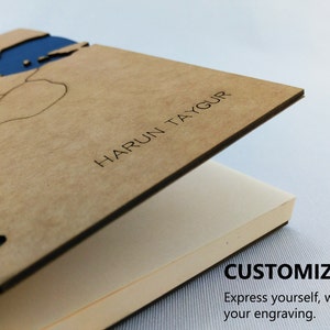 May include: A brown wooden notebook with a blue map design on the cover. The cover is open to reveal blank pages. The notebook is personalized with the name "HARUN TAYGUR".