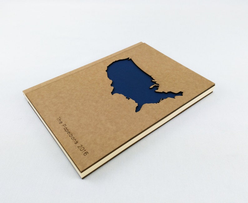 Puede incluir: Una libreta marr&oacute;n con una forma recortada de los Estados Unidos de Am&eacute;rica. El recorte est&aacute; relleno con un papel azul oscuro. La libreta tiene el texto "The Paperbons 2016" impreso en la portada.