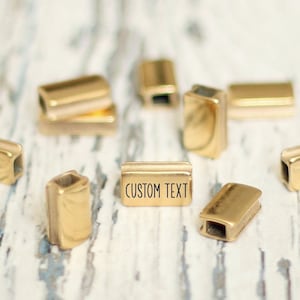 Könnte beinhalten: Goldfarbene Metallperlen in rechteckiger Form. Eine Perle trägt die Gravur "CUSTOM TEXT".