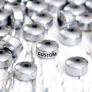 May include: A collection of shiny, silver-colored metal beads. Each bead has a flat, circular shape with a central hole. One bead is labeled with the word "CUSTOM" in black text. The beads are scattered on a white, textured surface.